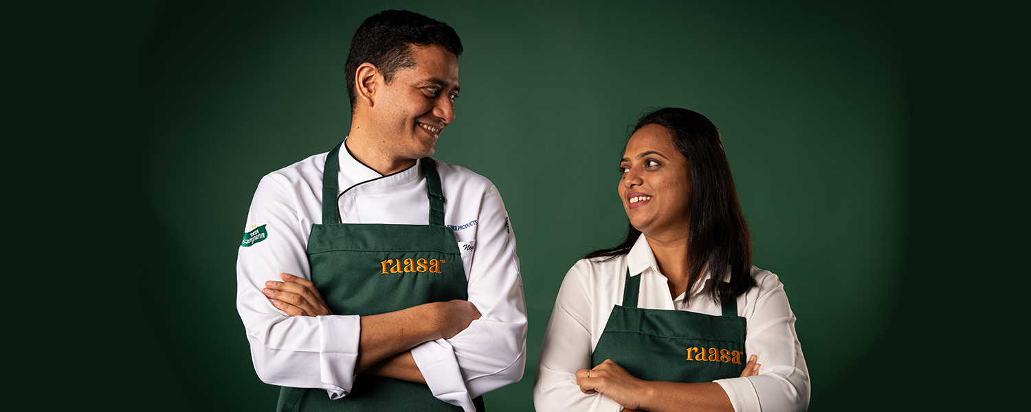 A picture of Chef Monali and Chef Yogi, the minds behind the decadent Authentic Indian meals that Raasa offers. They are both smiling, looking at each other. Chef Yogi is wearing a chef;s jacket and Monali is wearing a white shirt. They are both wearing a dark green apron with the word Raasa embroidered