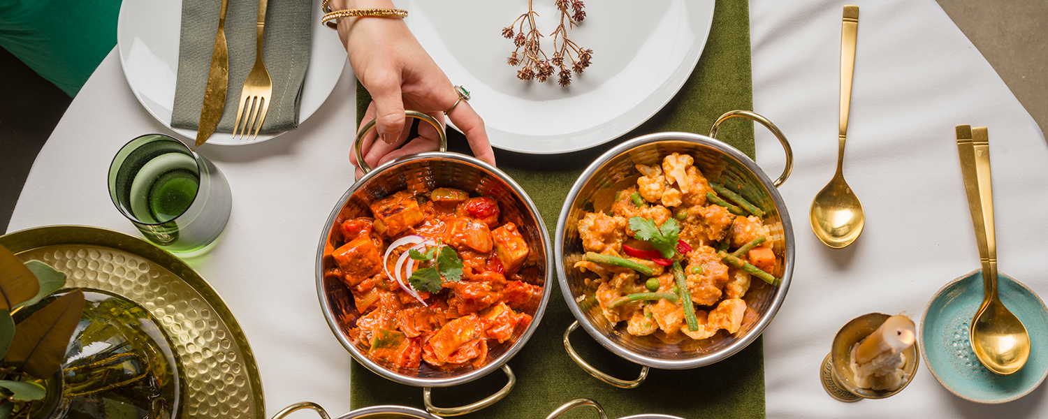 A picture if two gold plates , each holding one of the Raasa Authentic Indian Curries. The background shows it is a table set , with a dark green table cloth and gold cutlery and candles. There is a hand with gold dangles and rings, reaching for the Raasa Regal Tikka Masala curry
