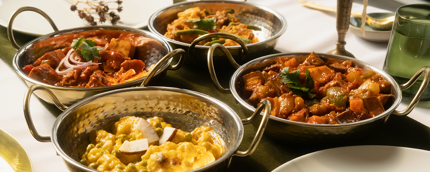 A picture from the side of four gold plates, close up. These hold each of the authentic Raasa Indian curries. A creamy Korma, A rich sambar, an authentic Kadai curry and delicious tikka masala 
