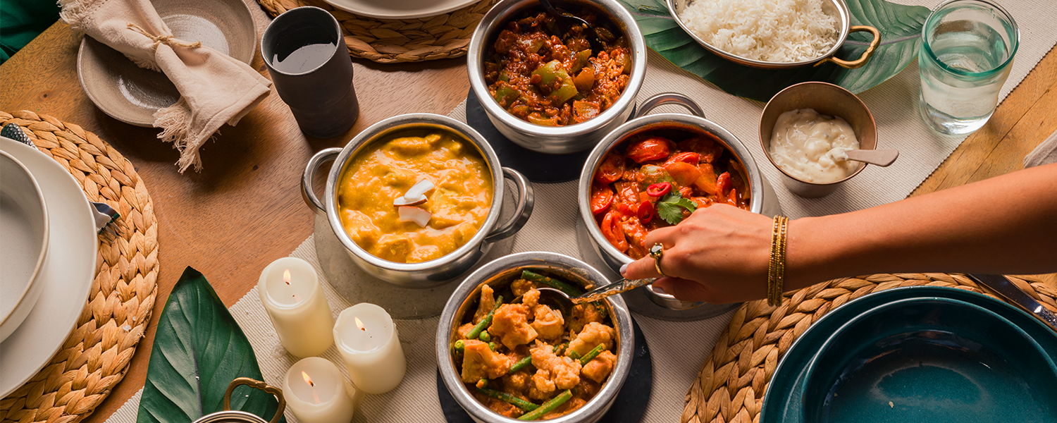 A picture of a table set with four Raasa curries. In the centre, the four dishes, Regal Tikka Masala, Royal Korma, Rich Sambar, and Majestic Kadai Curry. There is a hand coming from the right side reaching for the regal authentic Tikka Masala. The hand has beautiful gold jewelry. On the sides of the table, there are banana leaves adorning the table with plates of rice on top of them. 