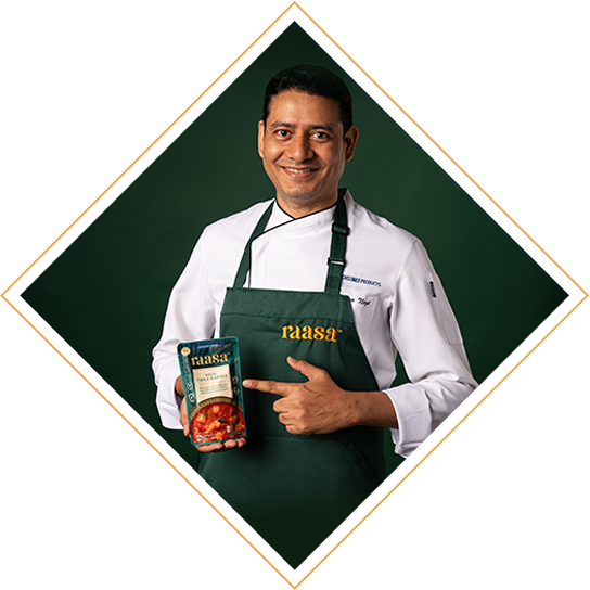 A picture cropped in the shape of a diamond. This is Chef Yogi, the Raasa chef from India. He is wearing a chefs jacket and a dark green apron that has the word Raasa embroidered in gold. He is holding a pack of Raasa Regal Tikka masal sauce and smiling widely. The background is dark green