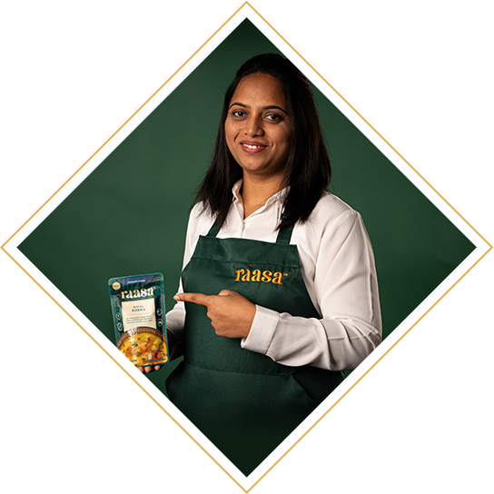A picture cropped in the shape of a diamond. This is Chef Monali, the Raasa food technologist and expert from India. She is wearing a chefs jacket and a dark green apron that has the word Raasa embroidered in gold. She is holding a pack of Raasa Royal Korma sauce and smiling widely. The background is dark green